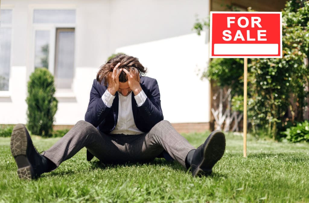 Depressed real estate agent sitting on grass in front of house for sale, outdoors